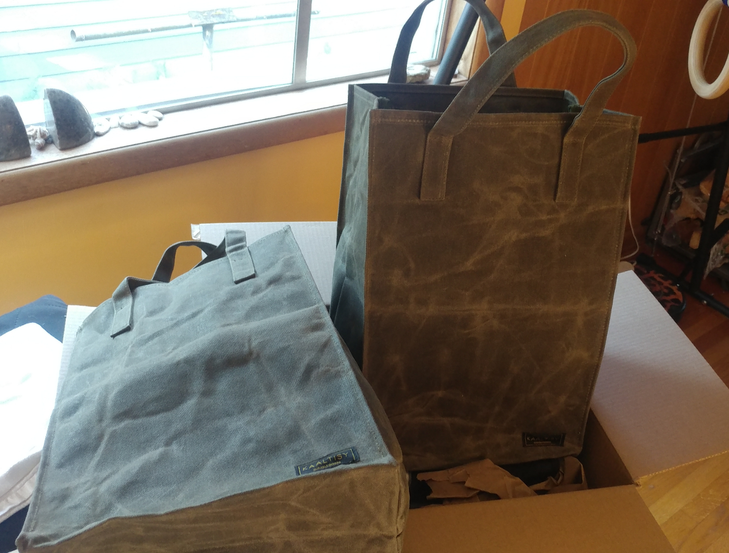 Two gray waxed canvas tote bags, one upright and one lying flat, are sitting on top of a cardboard box near a window with light coming in. The bags have short handles and stitched labels on the front.
