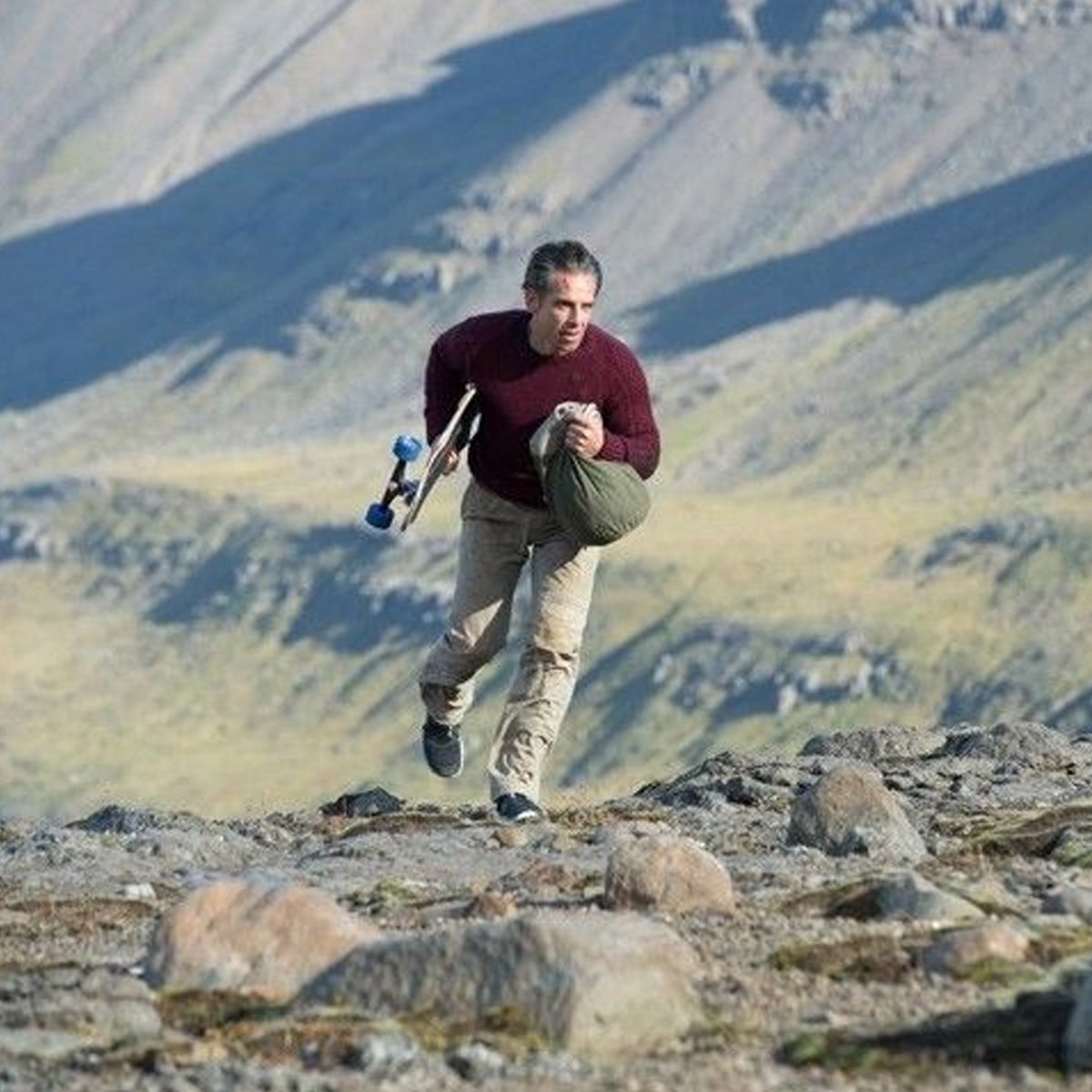 A man in a maroon sweater and beige pants carries a skateboard and a green duffel bag while walking over rocky terrain with hills and mountains in the background.