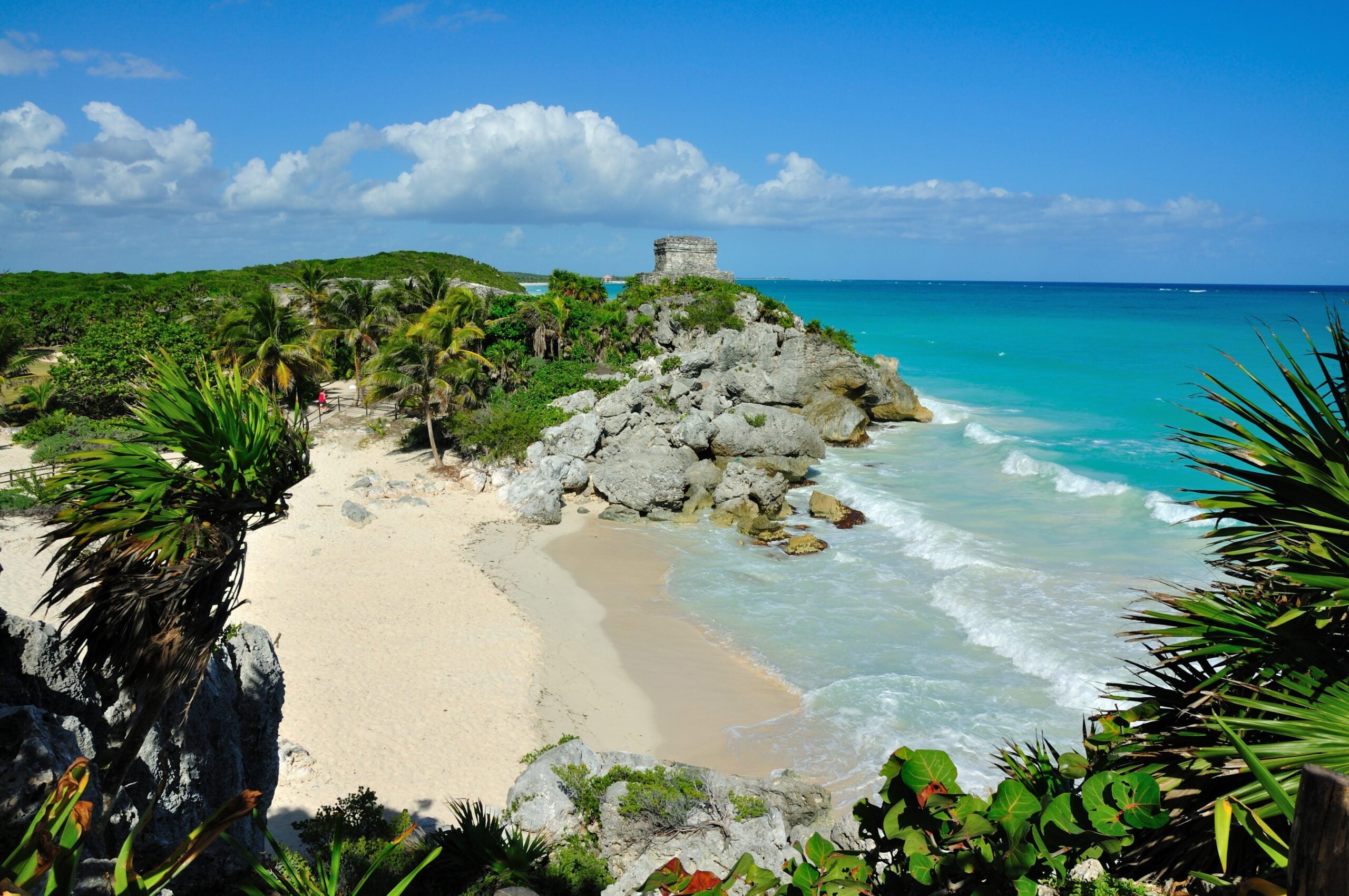 A sandy beach with turquoise waves, rocky cliffs, and lush green palm trees. An ancient stone structure sits atop the cliff, overlooking the Caribbean Sea under a bright blue sky with scattered clouds.