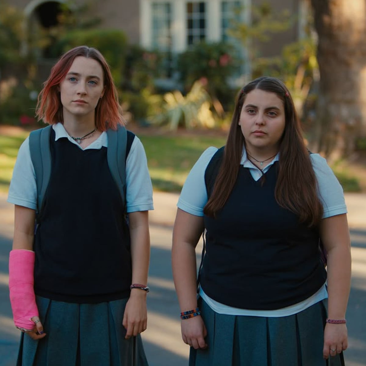 Two teenage girls in school uniforms stand side by side outdoors; one has short, reddish hair and a pink cast on her arm, while the other has long brown hair. Both have neutral expressions.