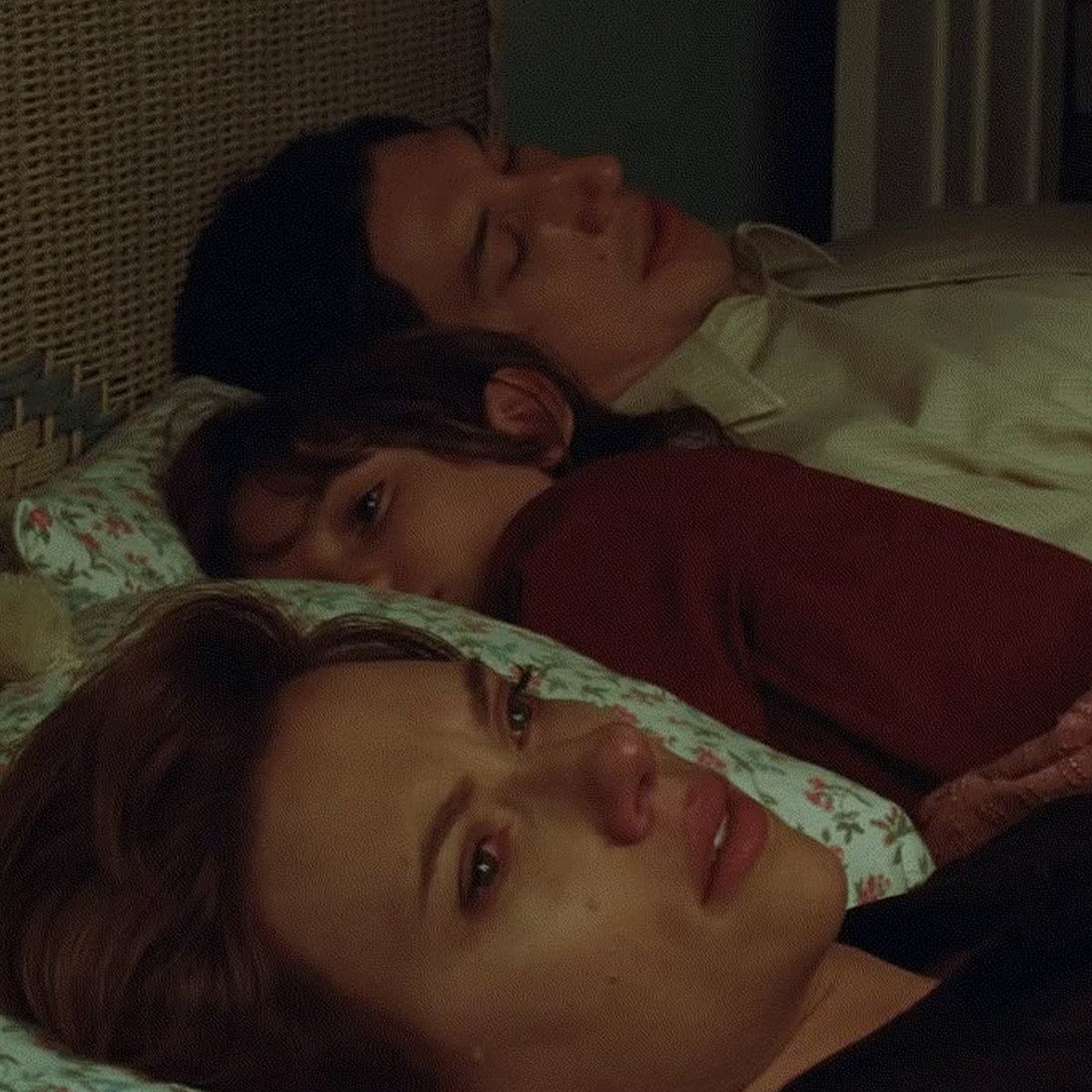 A woman lies in bed crying with tears on her face, while a man and a child sleep beside her in the background. The scene appears emotional and somber, with soft lighting and patterned bedding.