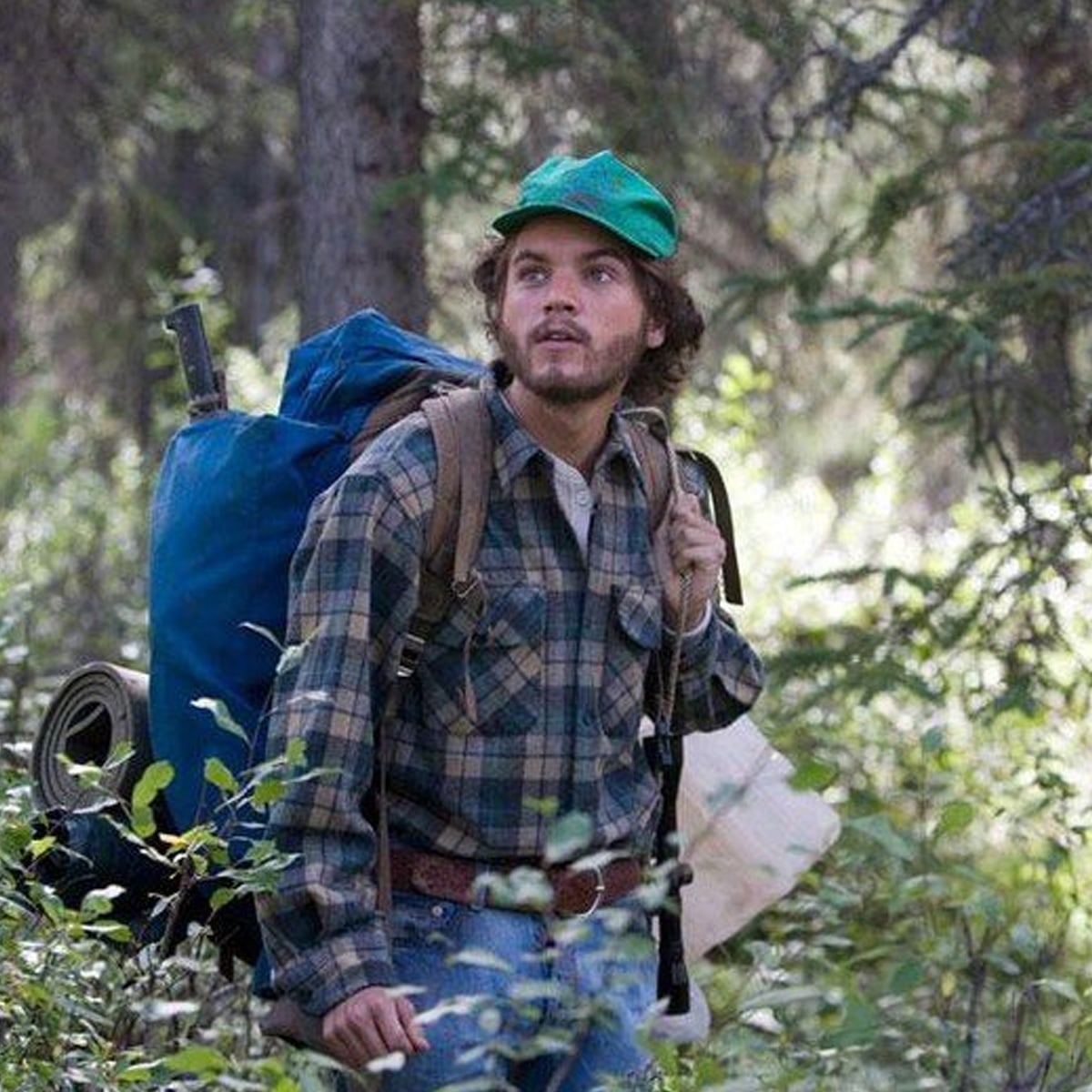 A man wearing a green cap and plaid shirt carries a large blue backpack while hiking through a dense forest, surrounded by trees and greenery.