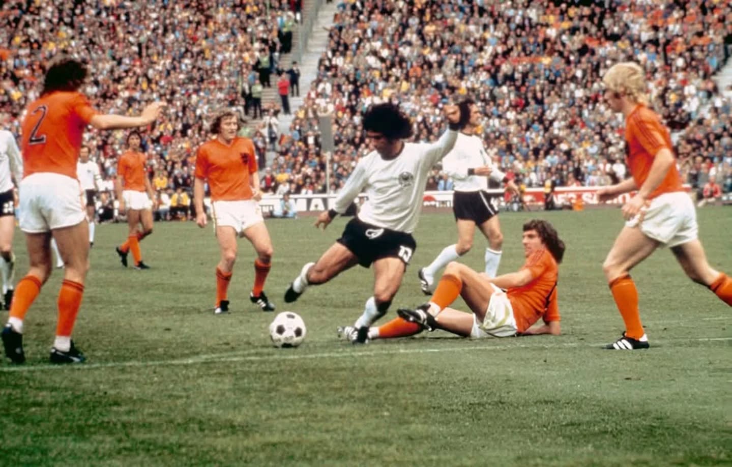 A soccer player in a white jersey is tackled by a player in an orange jersey during a crowded match, with several other orange-jerseyed players nearby and a stadium full of spectators in the background.