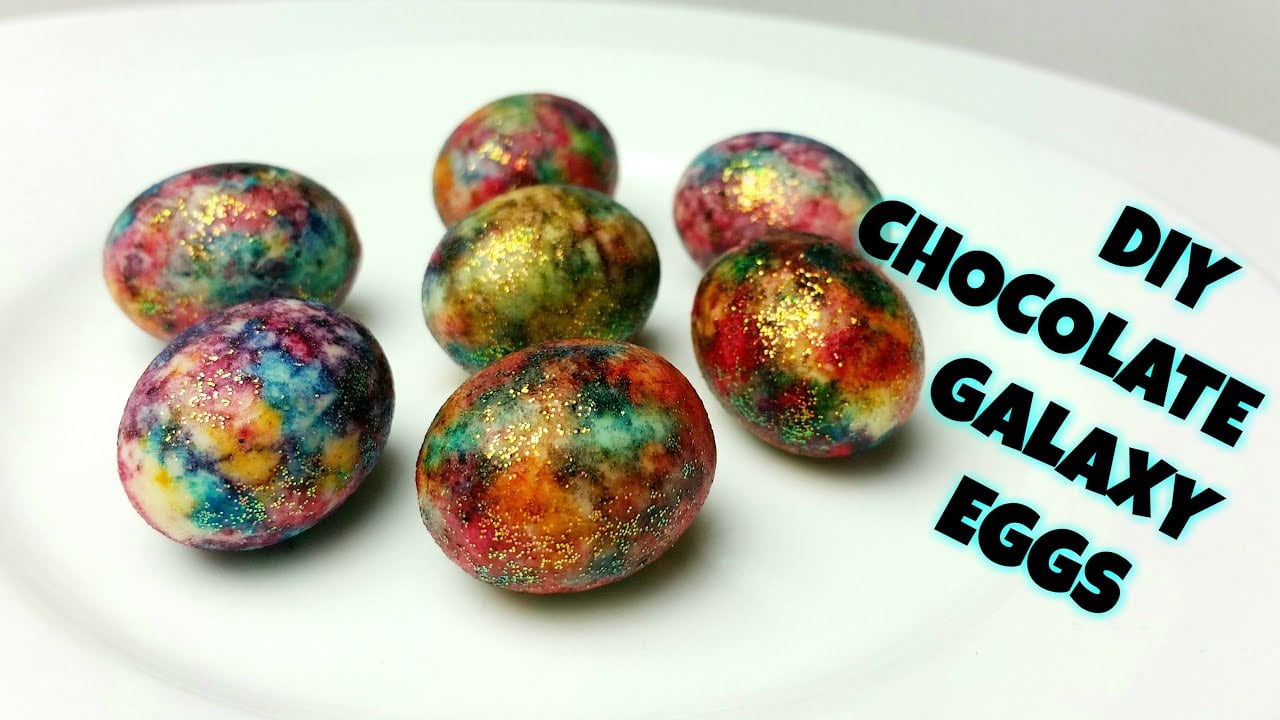 A white plate holds seven colorful, galaxy-patterned chocolate eggs with a shiny, glittery finish. The text on the right reads: "DIY Chocolate Galaxy Eggs" in bold, black letters.