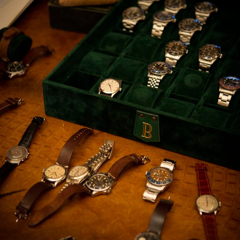 A green velvet watch box with a gold "B" holds several metal wristwatches, while various vintage watches with leather and metal bands are displayed on a brown textured surface nearby.