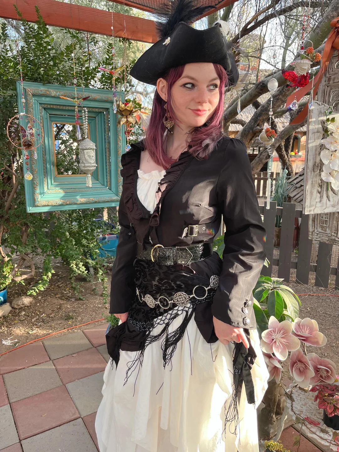 A person dressed in a pirate costume with a black hat, black jacket, white ruffled shirt, and a layered belt stands outdoors among plants, flowers, and decorative frames in bright sunlight.
