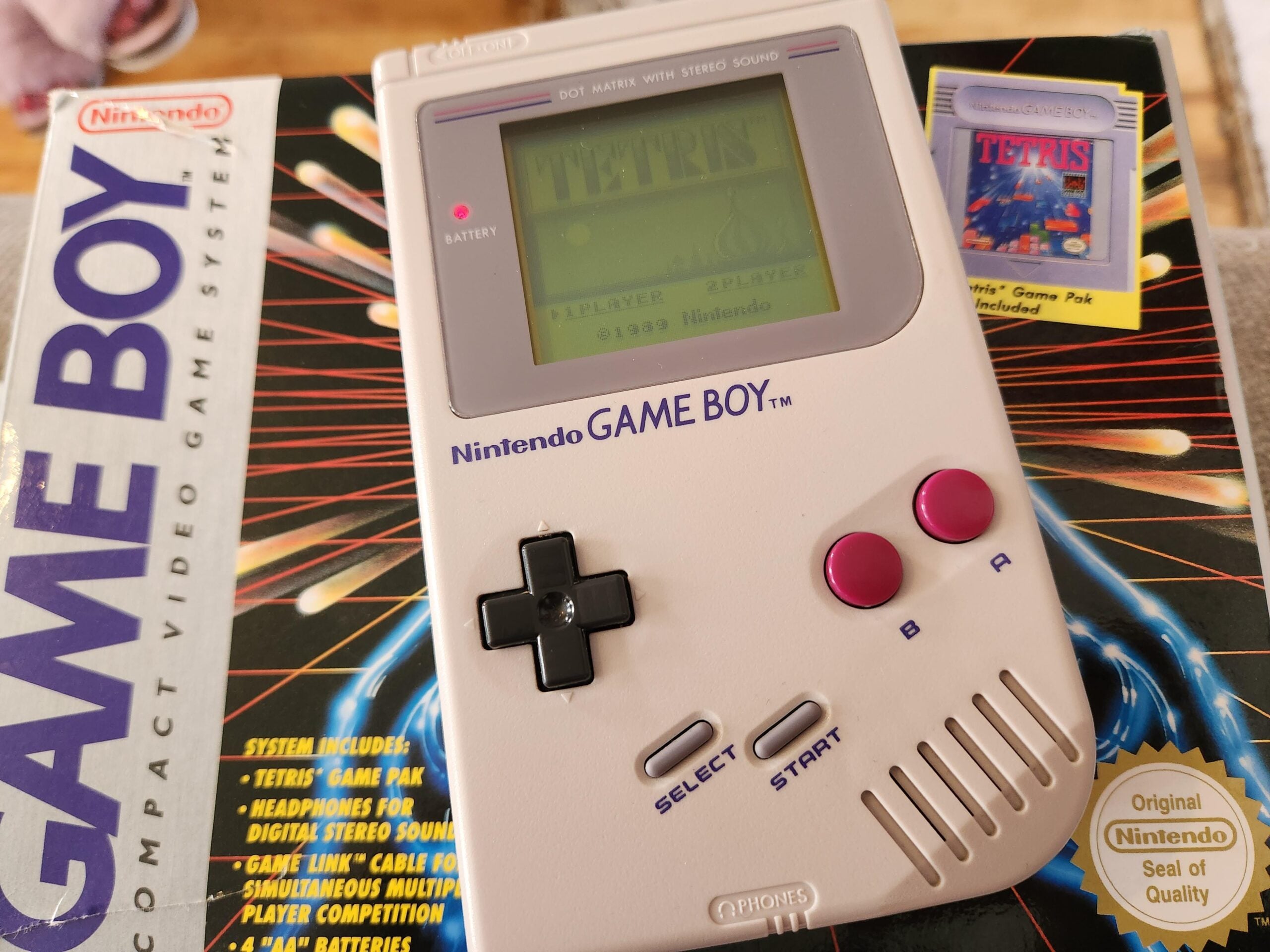 A Nintendo Game Boy displaying the game Tetris sits on top of its original box, which also shows a Tetris game pack. The box features colorful graphics and lists features of the Game Boy system.