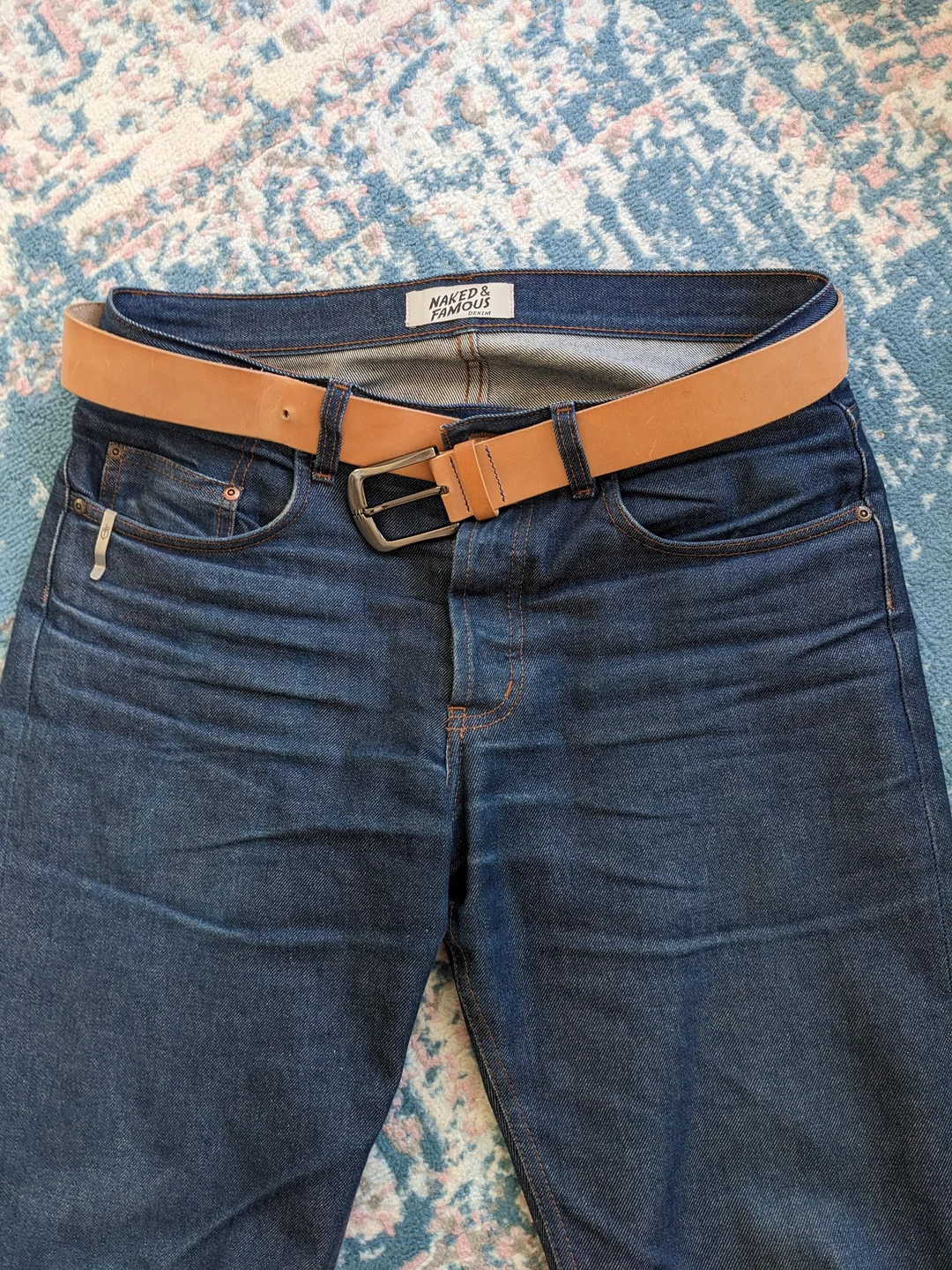A pair of dark blue jeans with a light brown leather belt threaded through the loops, lying flat on a blue and pink patterned surface.