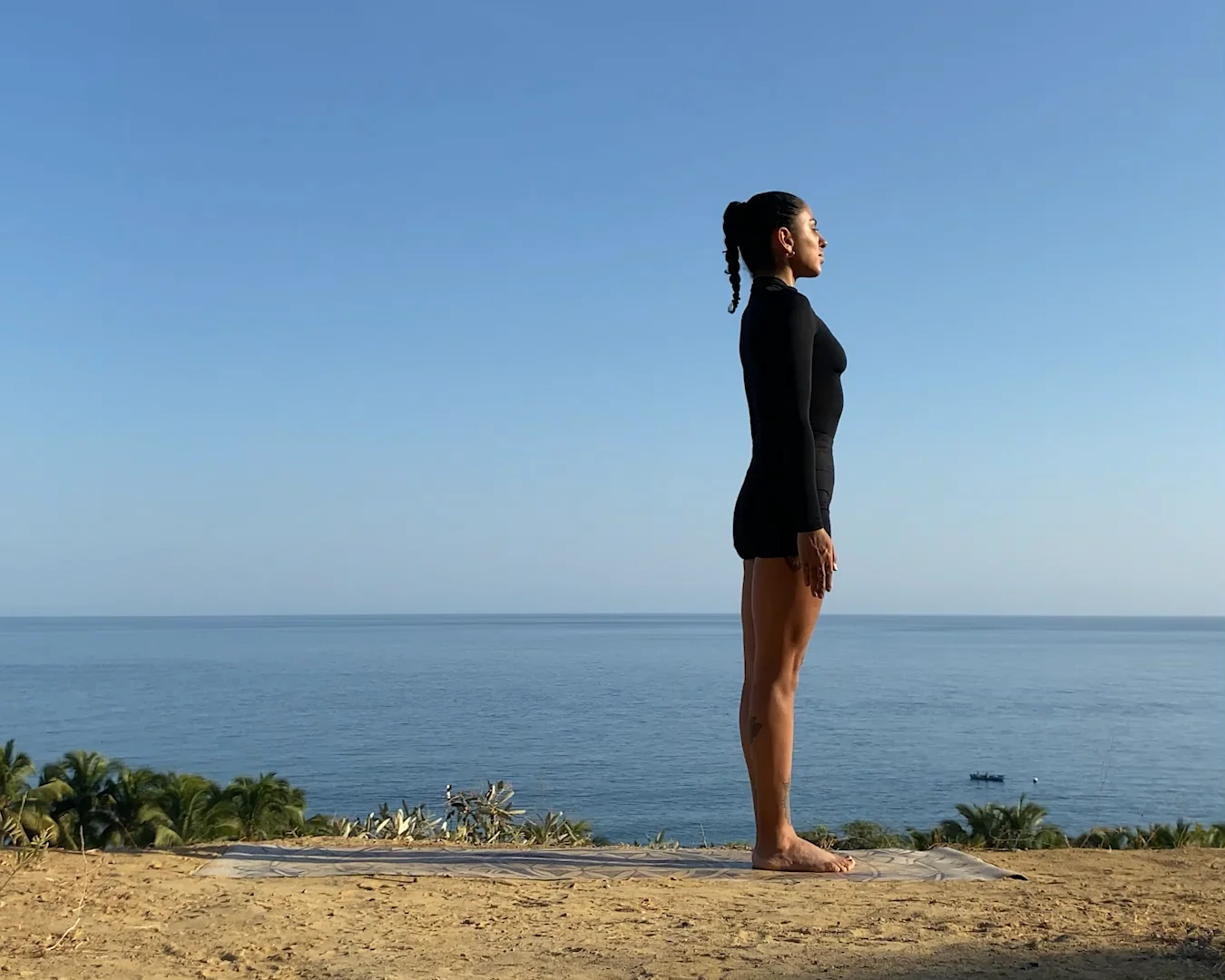 A woman stands in a yoga pose on a mat outdoors, near the edge of a sandy surface overlooking the ocean, with clear blue sky and greenery in the background.