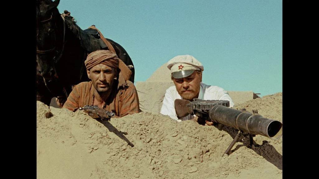 Two men lie in a sandy trench aiming rifles; one wears a brown headscarf and the other a white Soviet military cap. There is a black horse behind them and clear blue sky overhead.