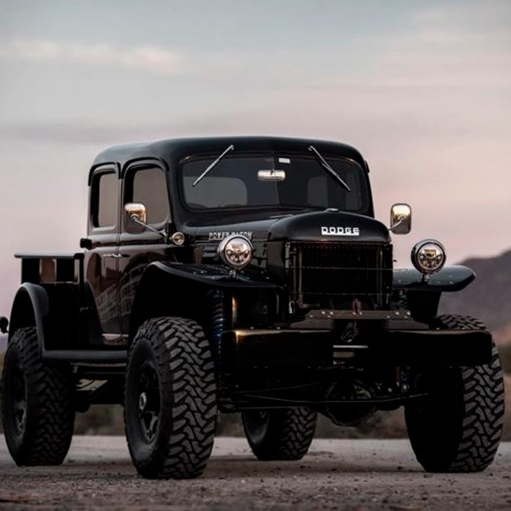 A restored black vintage Dodge Power Wagon pickup truck with large off-road tires is parked outdoors on a dirt road, with mountains and a cloudy sky in the background.