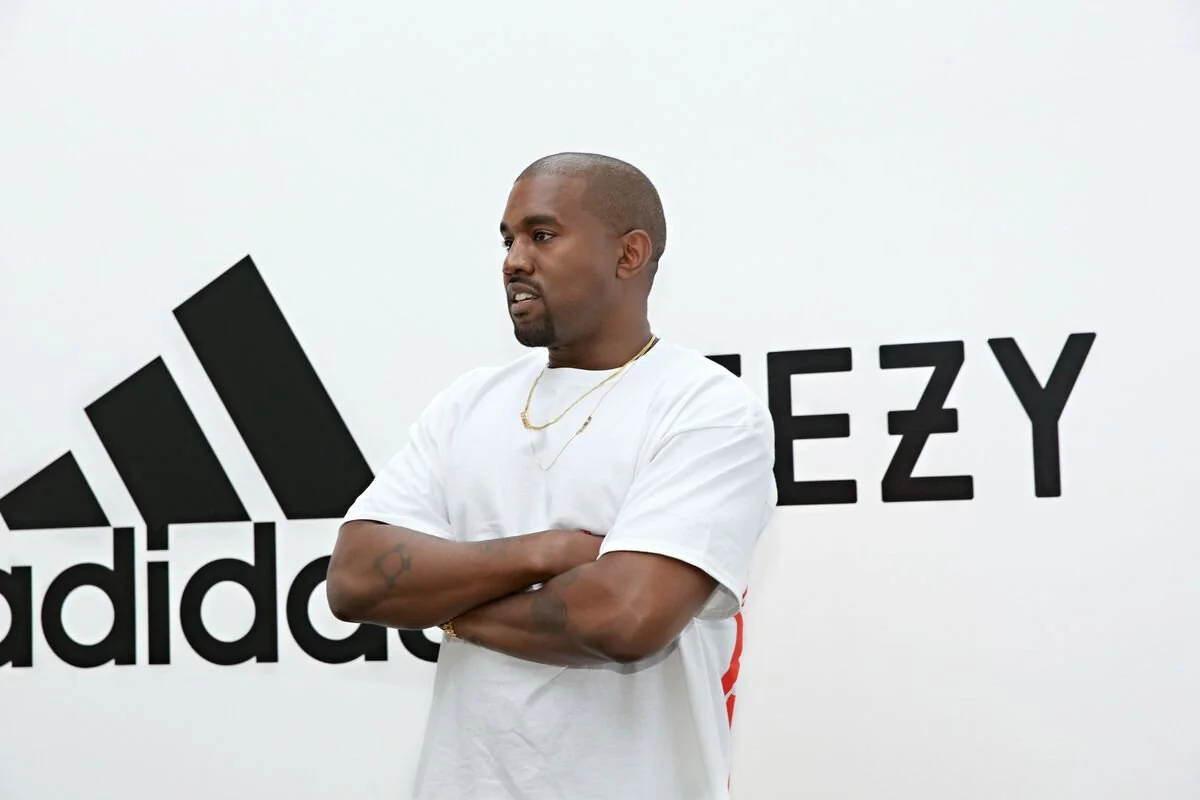 A man in a white t-shirt stands with his arms crossed in front of a wall with the Adidas logo and the word “YEEZY.”