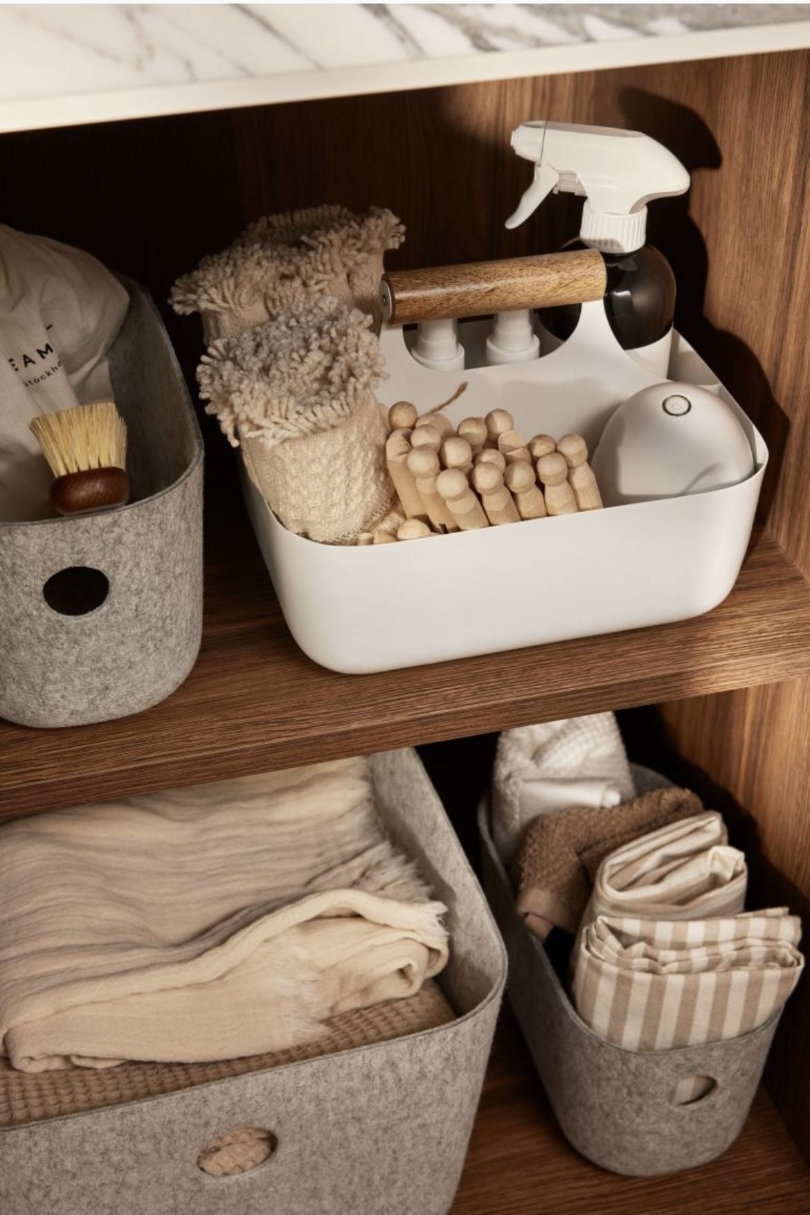 White and gray storage bins on wooden shelves hold cleaning tools, towels, scrub brushes, cloths, a spray bottle, and wooden laundry pins, all neatly organized for easy access.