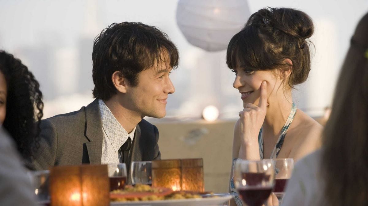 A man and a woman sit at an outdoor table, smiling and gazing at each other. The table has drinks and food, and there are candles and a paper lantern in the background, suggesting a romantic setting.