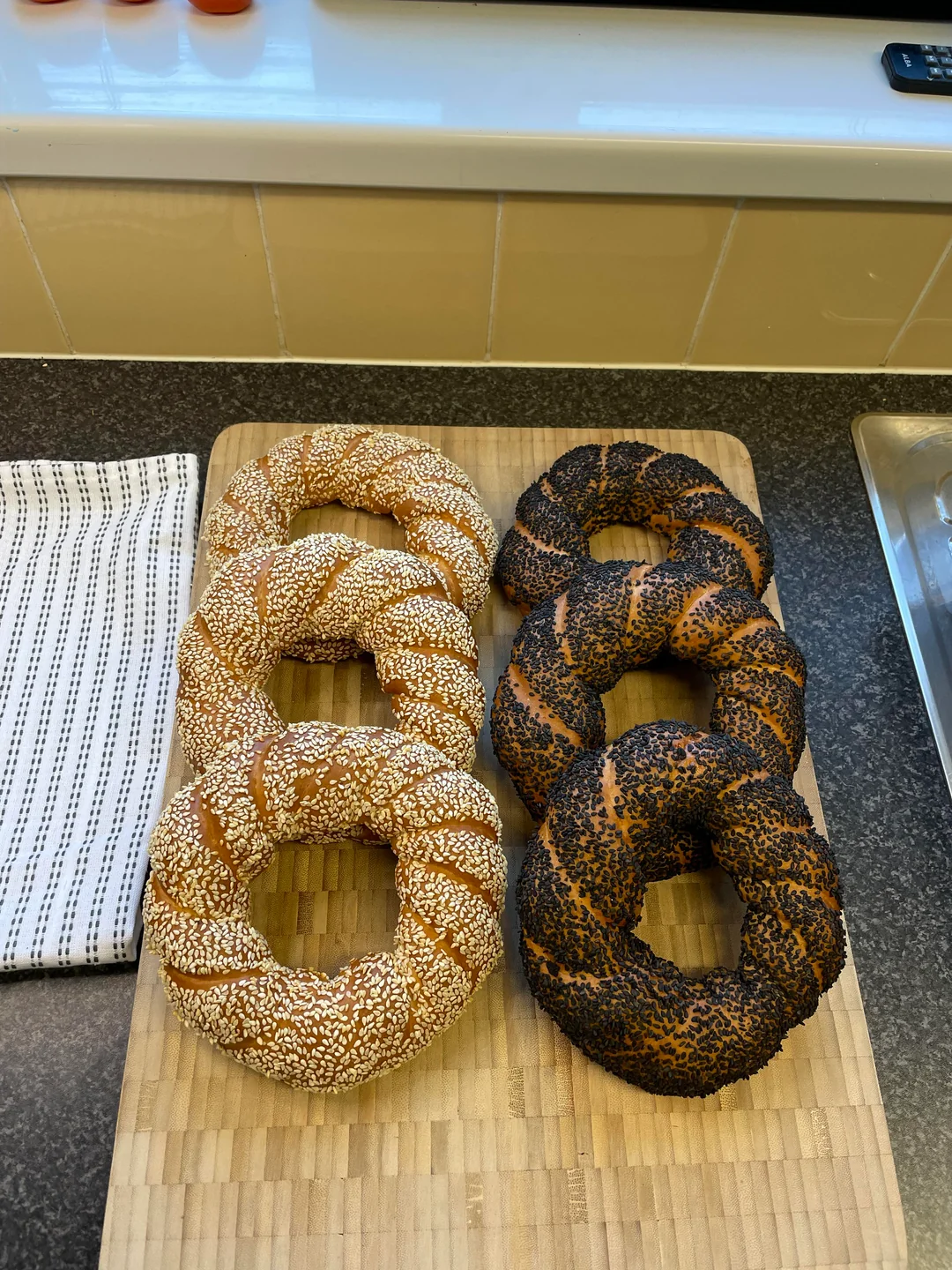 Six round braided breads are arranged in two columns on a wooden board: three topped with sesame seeds on the left and three with dark poppy seeds on the right. A striped towel sits nearby on the counter.