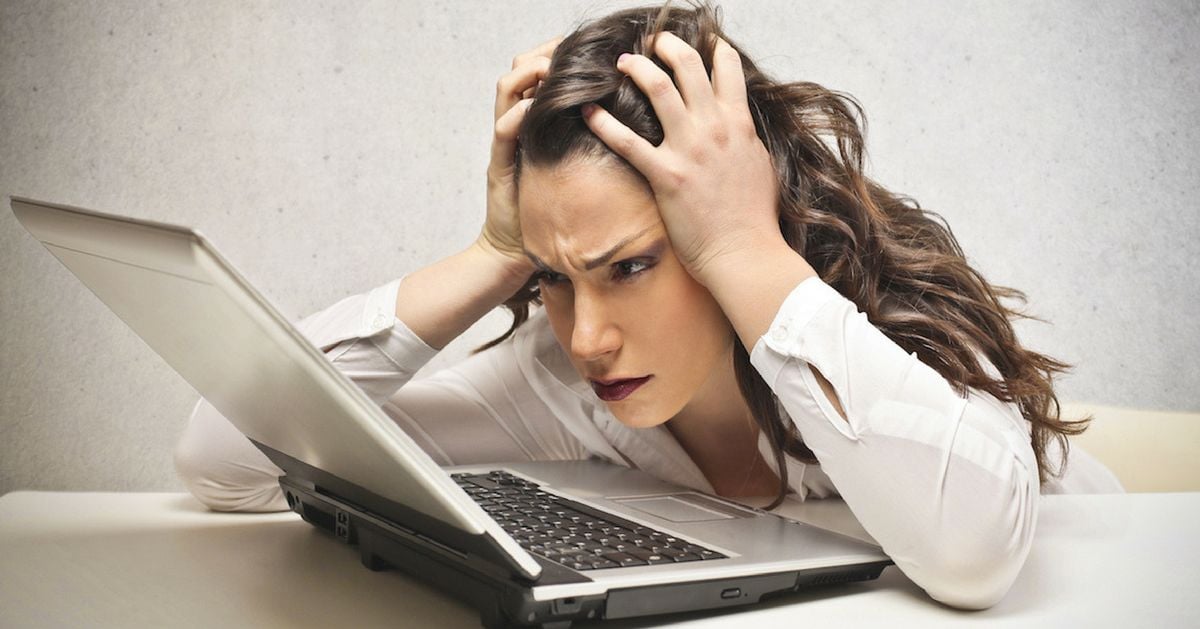 A woman with long, brown hair sits at a table, looking frustrated and stressed while holding her head in her hands and staring intently at an open laptop in front of her.