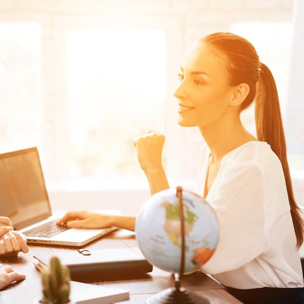A woman with a ponytail sits at a desk with a laptop, notebook, and small globe, smiling and talking to someone off-camera in a sunlit office.