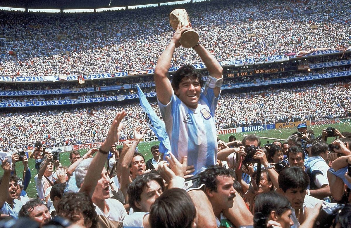 A jubilant soccer player in an Argentina jersey holds the World Cup trophy above his head while being lifted by celebrating teammates and fans in a packed stadium.