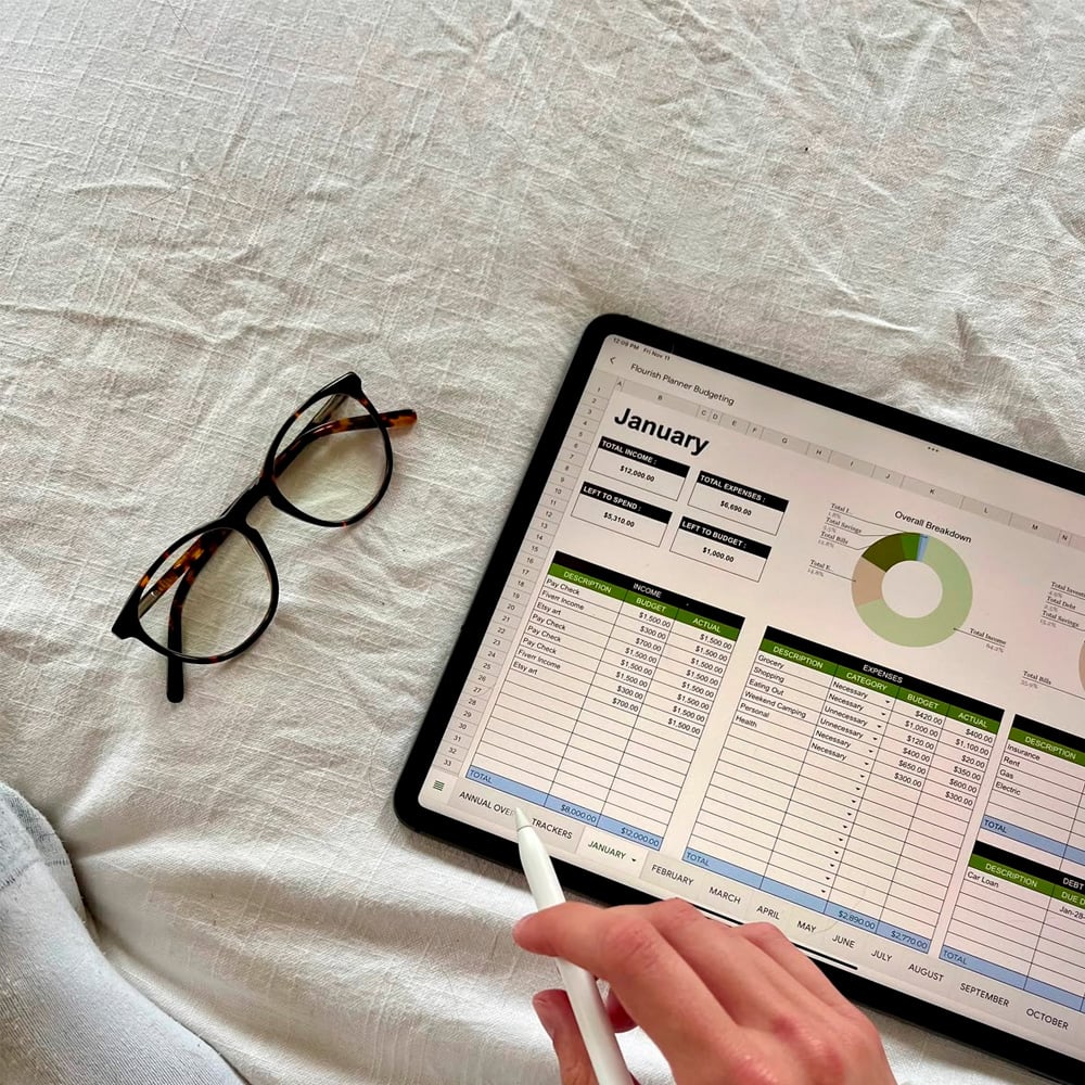 A person holds a stylus while pointing at a tablet screen displaying a financial spreadsheet for January; eyeglasses rest on a light fabric surface nearby.