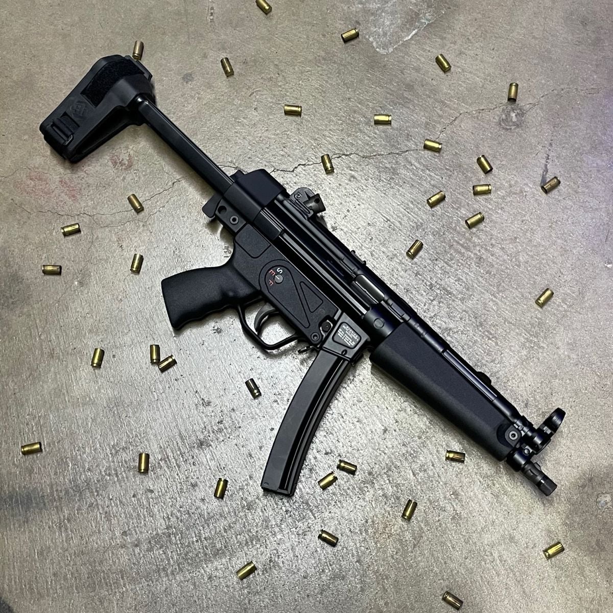 A black submachine gun with a folding stock and a curved magazine lies on a concrete floor, surrounded by scattered brass bullet casings.