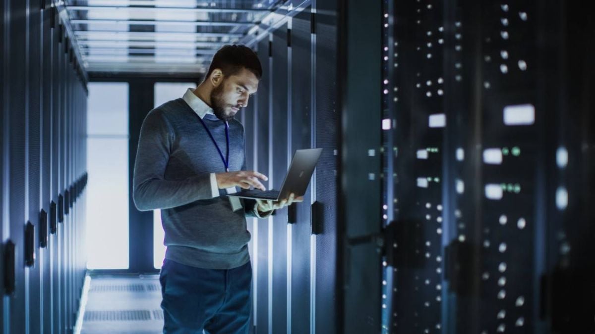 A man stands in a server room, working on a laptop. He is surrounded by tall racks of servers with blinking lights, suggesting he is performing IT or network maintenance.