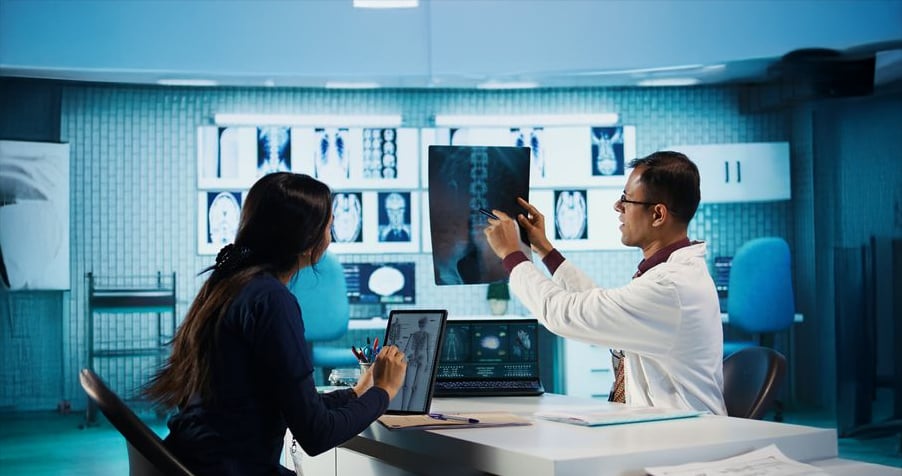 A doctor shows an X-ray image to a woman in a modern medical office. They sit at a desk with a laptop, clipboard, and notes, with illuminated medical scans displayed in the background.