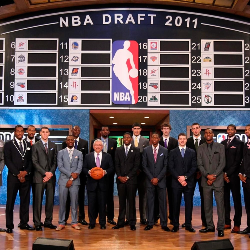 A group of suited young men and an older man holding a basketball stand on stage in front of an NBA Draft 2011 board featuring team logos and draft pick numbers.