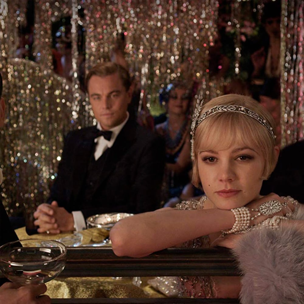 A woman in 1920s attire rests her arms on a table, looking pensive, while a man in a tuxedo sits behind her. Sparkling decorations and party guests fill the glamorous background.