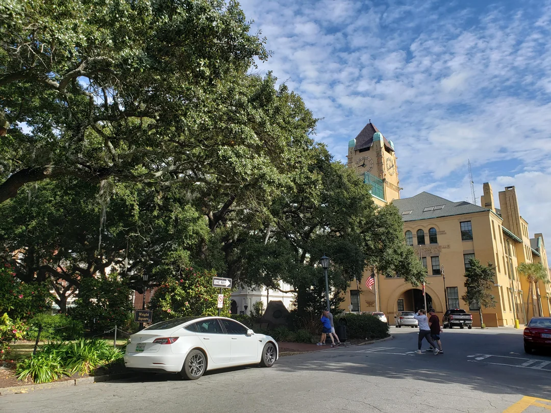 A white car is parked on a tree-lined street near a large yellow historic building with a clock tower. People walk on the sidewalk under a partly cloudy sky.