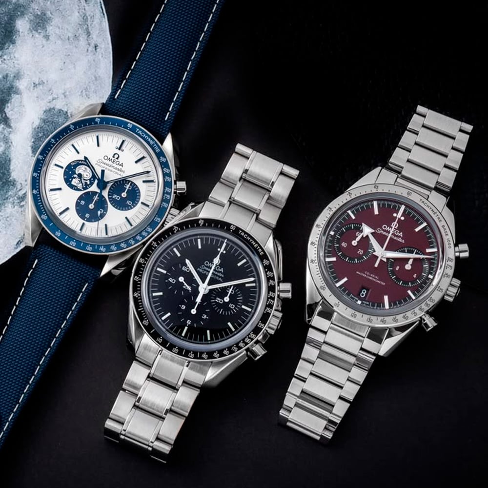 Three Omega Speedmaster watches are displayed side by side: one with a blue strap and white dial, one with a black dial and metal bracelet, and one with a burgundy dial and metal bracelet, against a dark background.