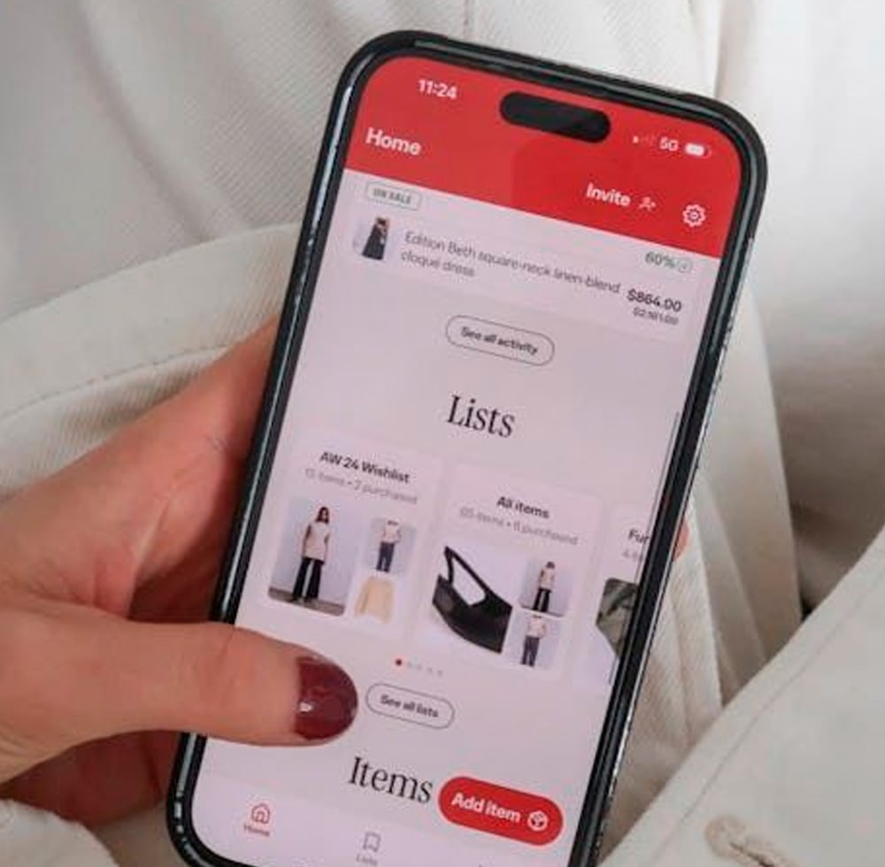 A person with red painted nails is holding a smartphone displaying a shopping app screen with clothing and accessory lists. The background shows a light-colored fabric, possibly clothing.