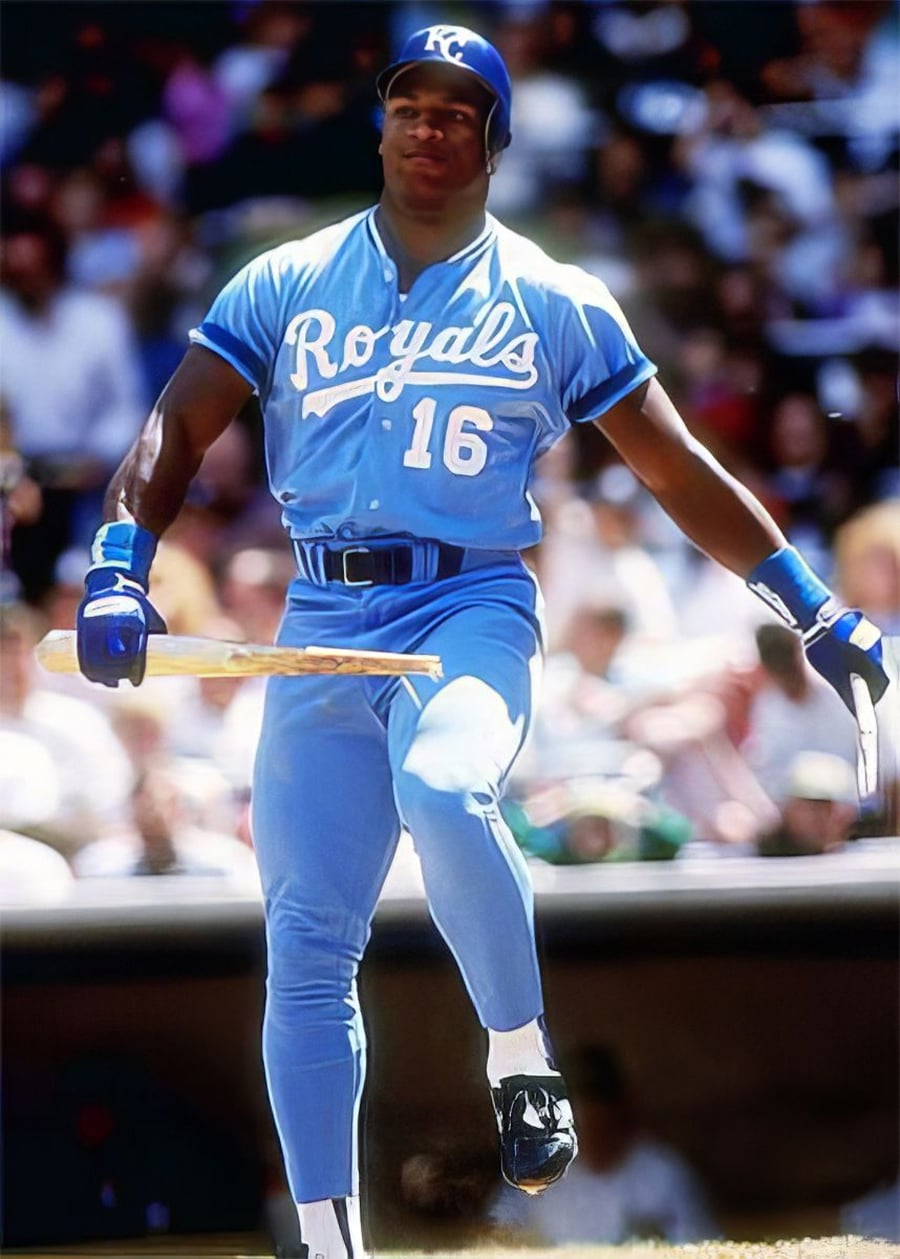 A baseball player in a blue Kansas City Royals uniform, number 16, breaks a bat while stepping forward on the field during a game, with spectators blurred in the background.
