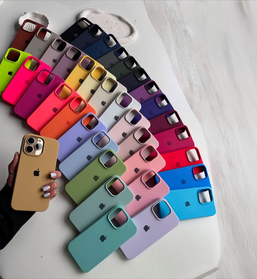A hand holds a beige iPhone with a matching case, surrounded by an array of colorful iPhone cases arranged in rows on a white surface. The cases are in various colors, including blue, pink, green, red, and more.