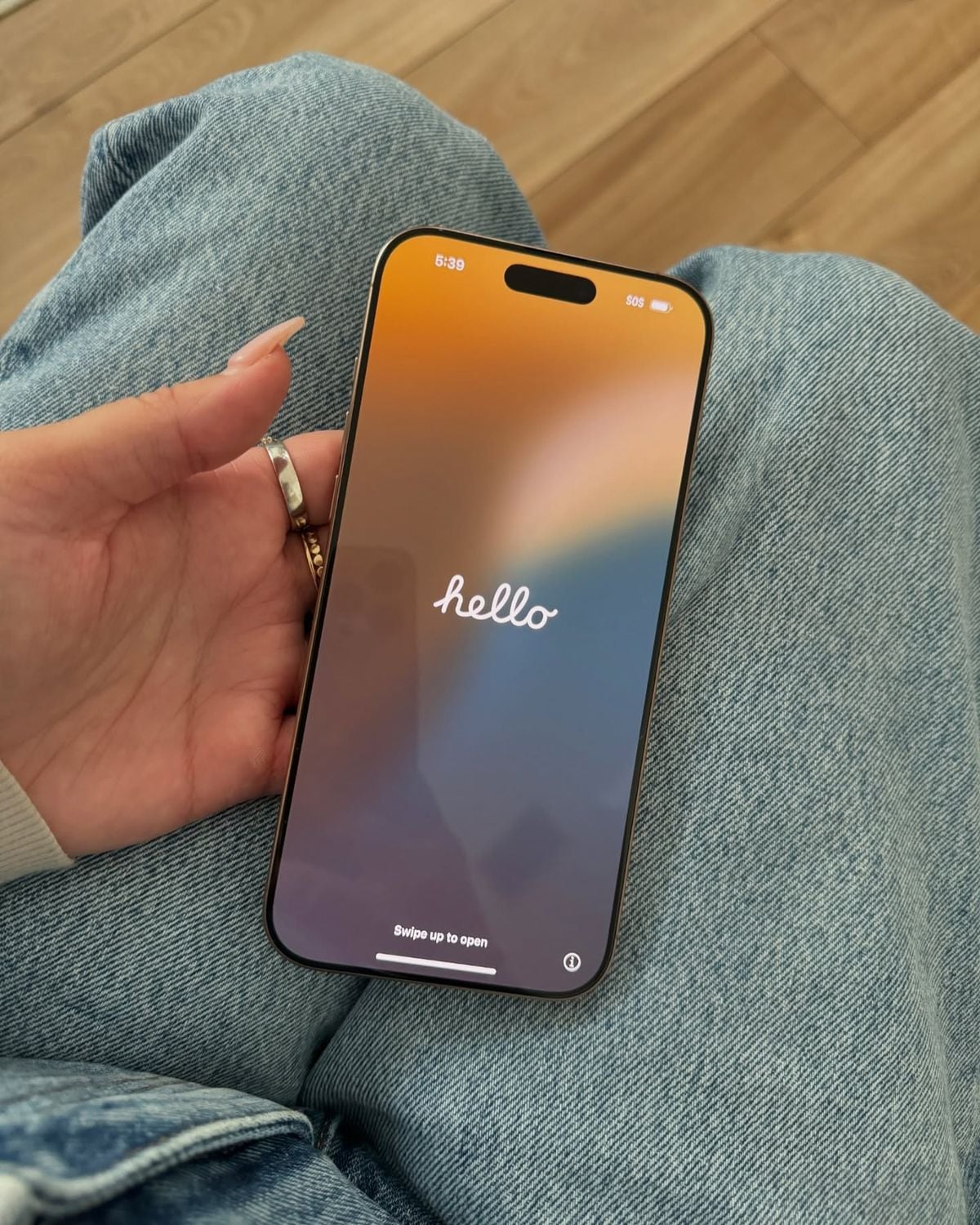A person holding a smartphone in their hand, resting on their denim-clad lap. The phone screen displays a colorful gradient background with the word "hello" in white text.