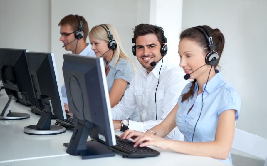 Four customer service representatives wearing headsets sit in a row at computers in a bright office. The person second from the right is smiling at the camera, while the others focus on their screens.
