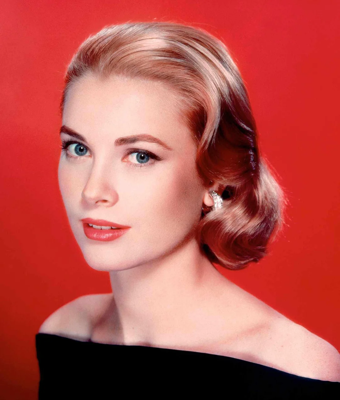 A woman with short, wavy blonde hair and blue eyes poses against a red background. She wears a black off-the-shoulder top, red lipstick, and pearl earrings, looking slightly to the side with a calm expression.