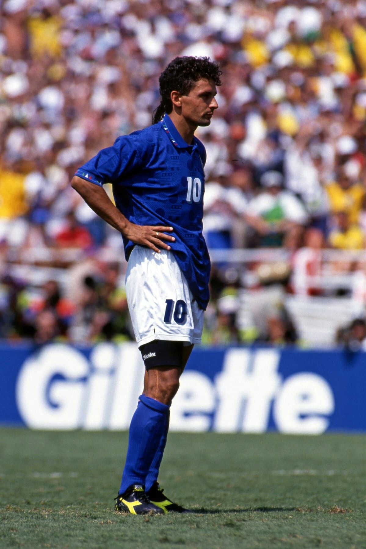 A male soccer player in a blue jersey with number 10 stands on a grassy field with his hands on his hips. A blurred crowd and a Gillette advertisement are visible in the background.