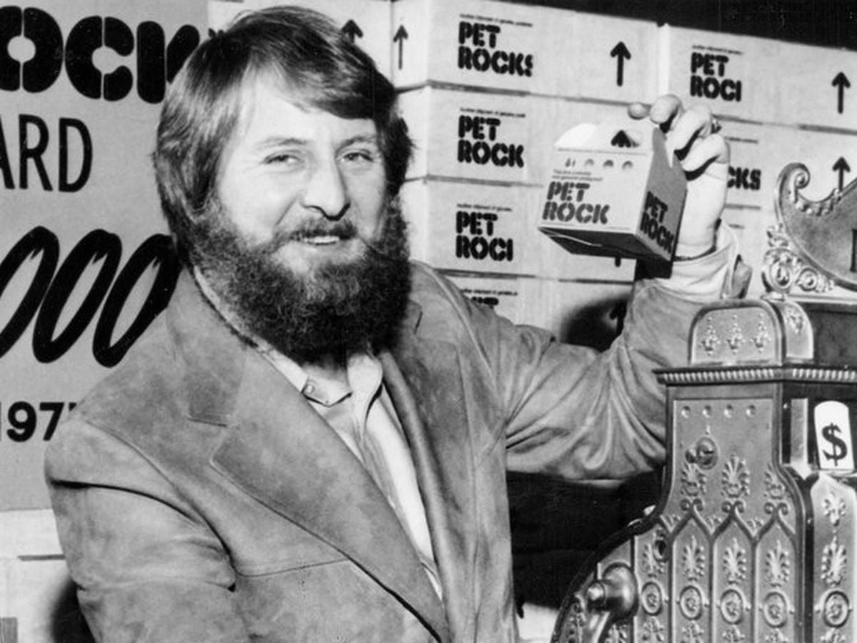 A bearded man in a suit smiles while holding a small box labeled "Pet Rock" next to an old-fashioned cash register, with boxes marked "Pet Rock" stacked in the background.