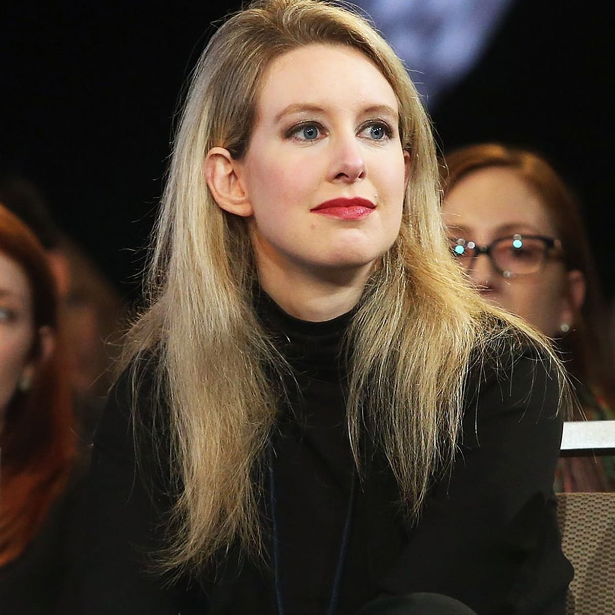 A woman with long blonde hair and fair skin sits among a group of people. She is wearing a black turtleneck and looking slightly to the side with a neutral expression. The background is out of focus.