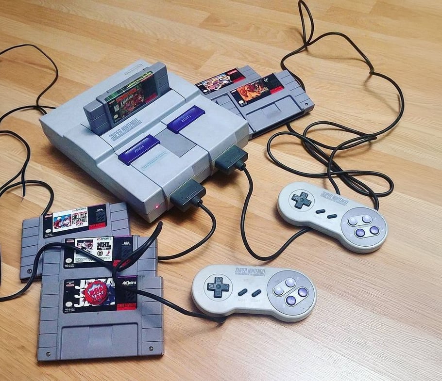 A Super Nintendo Entertainment System (SNES) console with two controllers and three game cartridges sits on a wooden floor, with one cartridge inserted in the console and the other two lying nearby.