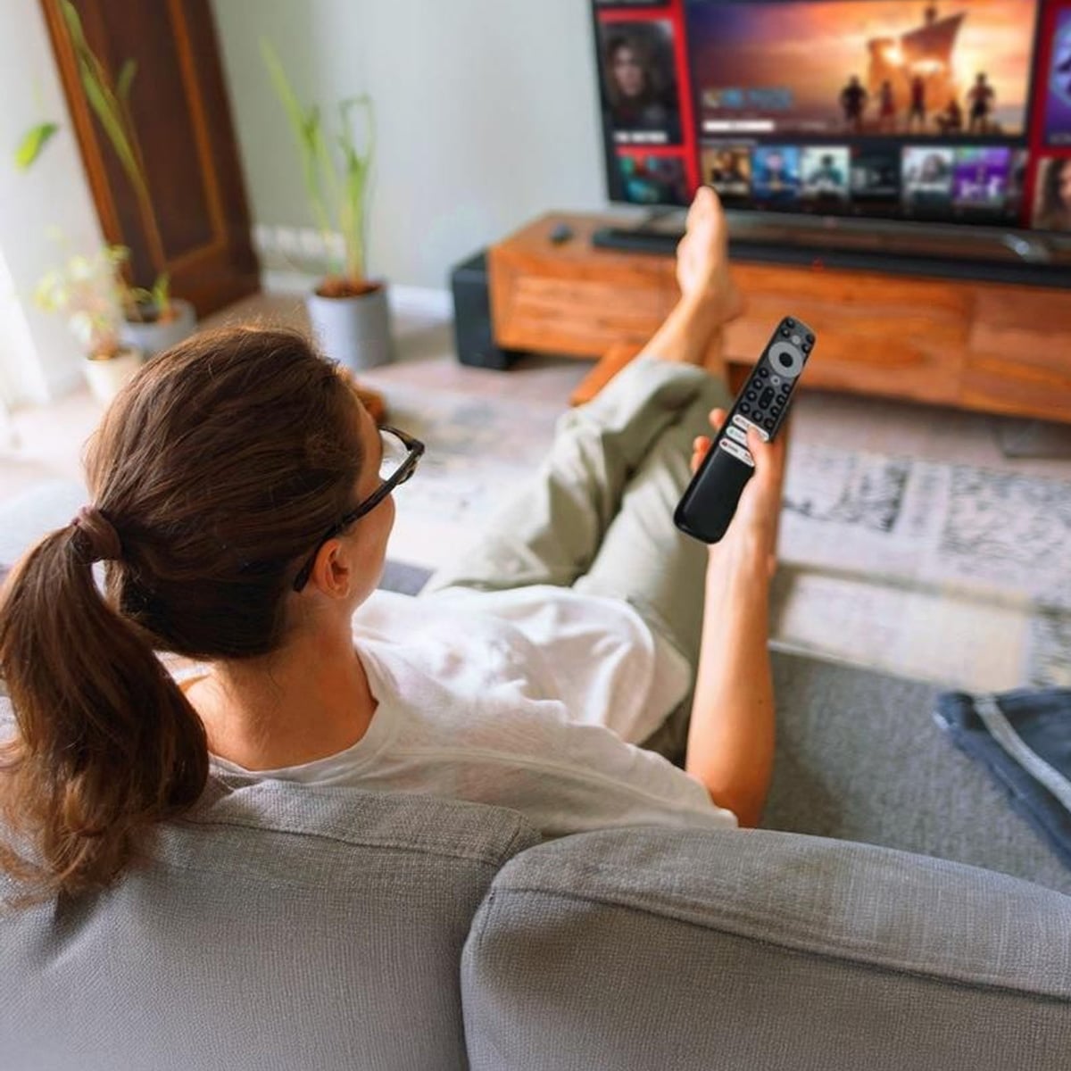 A person with brown hair in a ponytail sits on a couch, holding a TV remote and watching a streaming service on a flat-screen television in a modern living room.