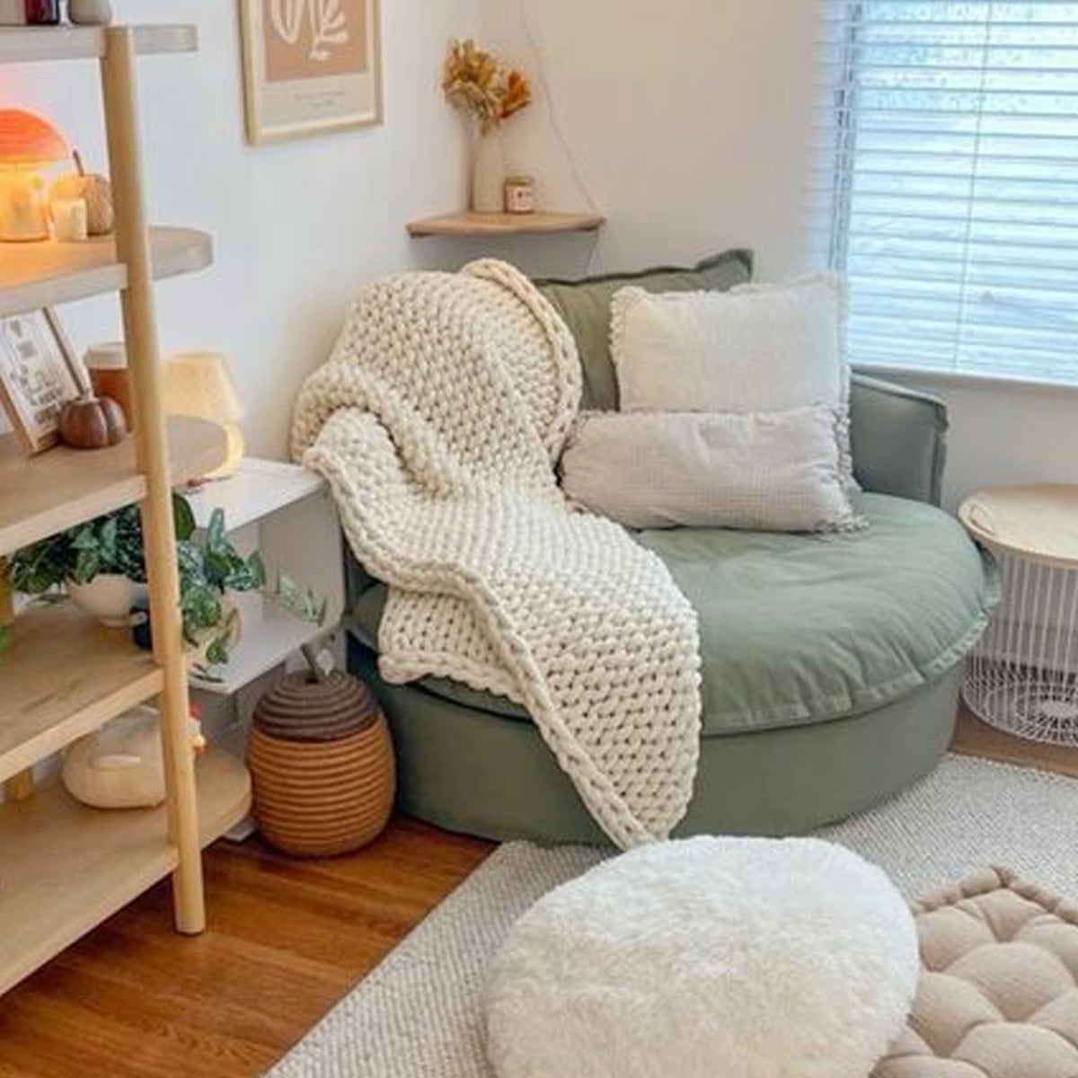 A cozy reading nook with a round green chair, layered with white cushions and a chunky knit blanket, sits by a window. Shelves with decor, a plant, and soft lighting create a warm, inviting atmosphere.