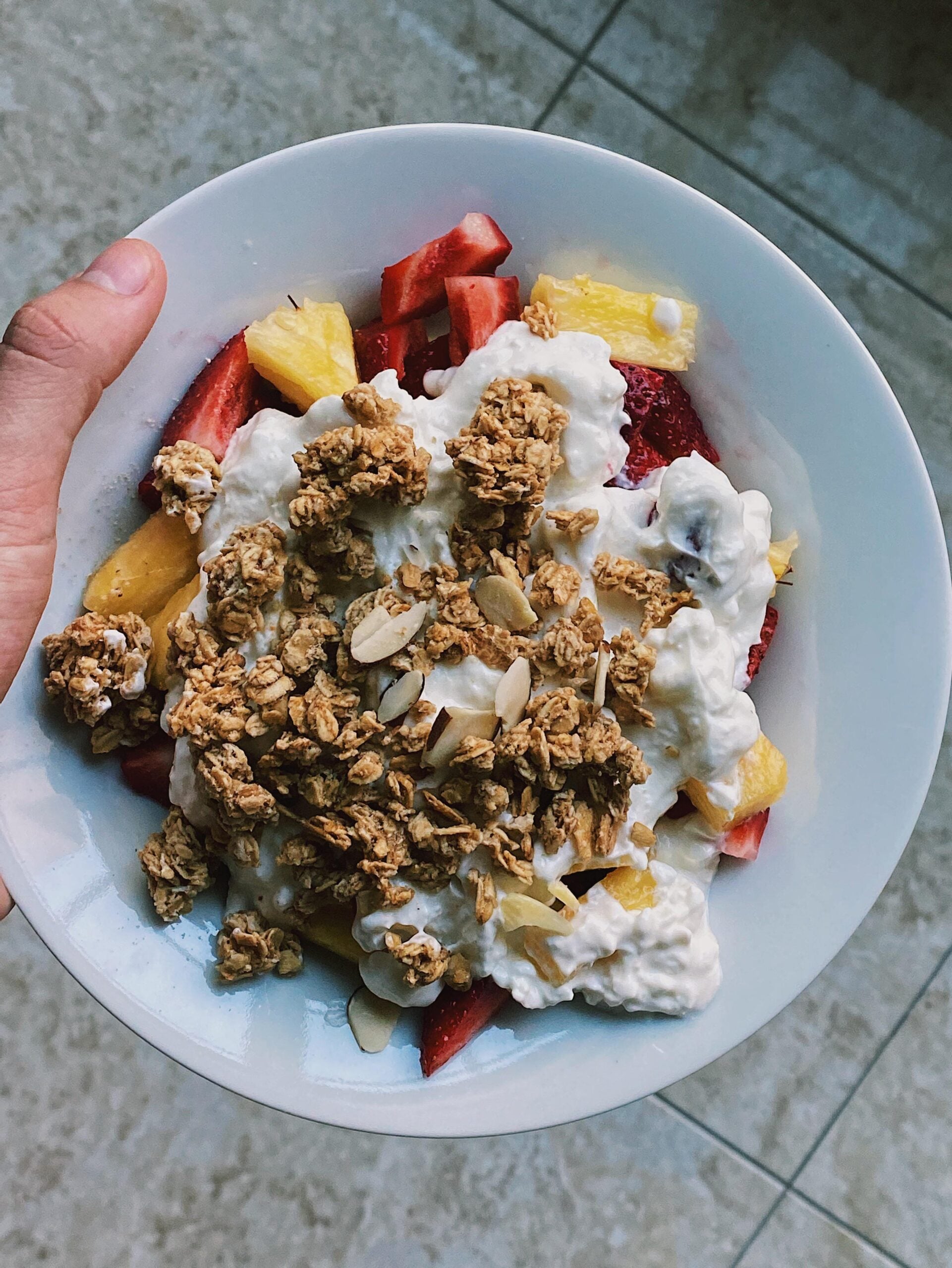 A hand holds a white bowl filled with strawberries, pineapple chunks, cottage cheese, granola, and sliced almonds, on a tiled floor background.