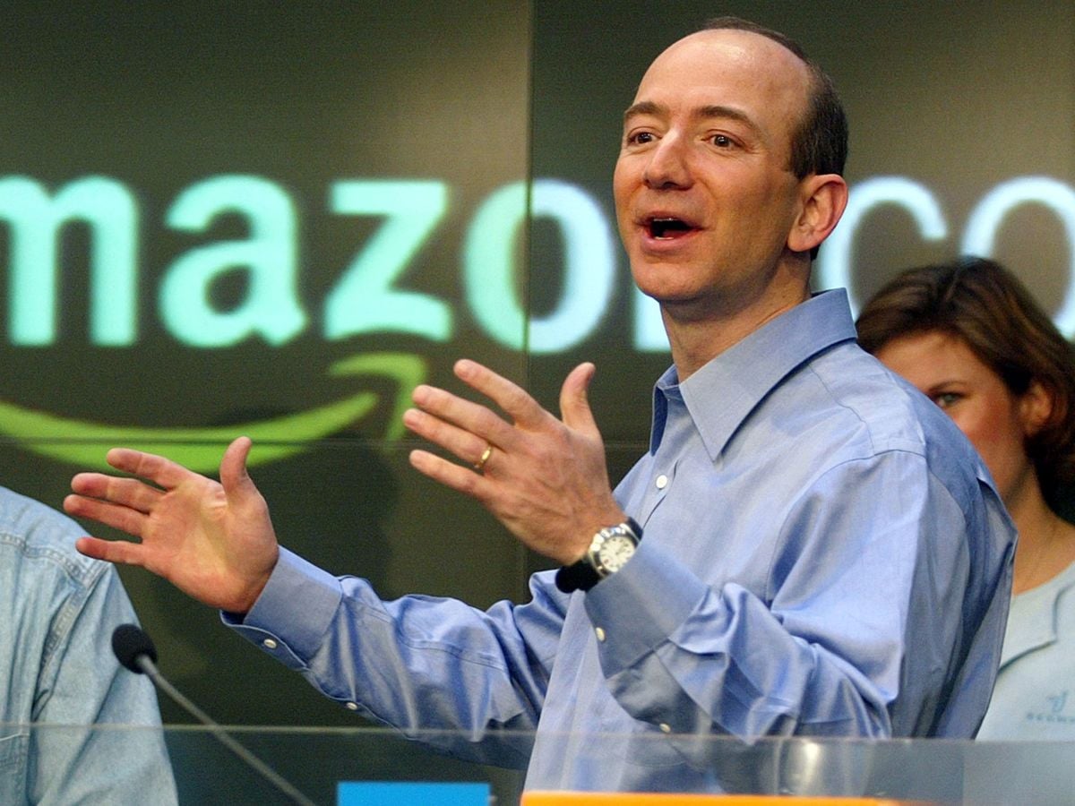 A man in a blue shirt speaks enthusiastically at a podium, gesturing with his hands. The blurred Amazon logo is visible in the background, with other people partially visible nearby.