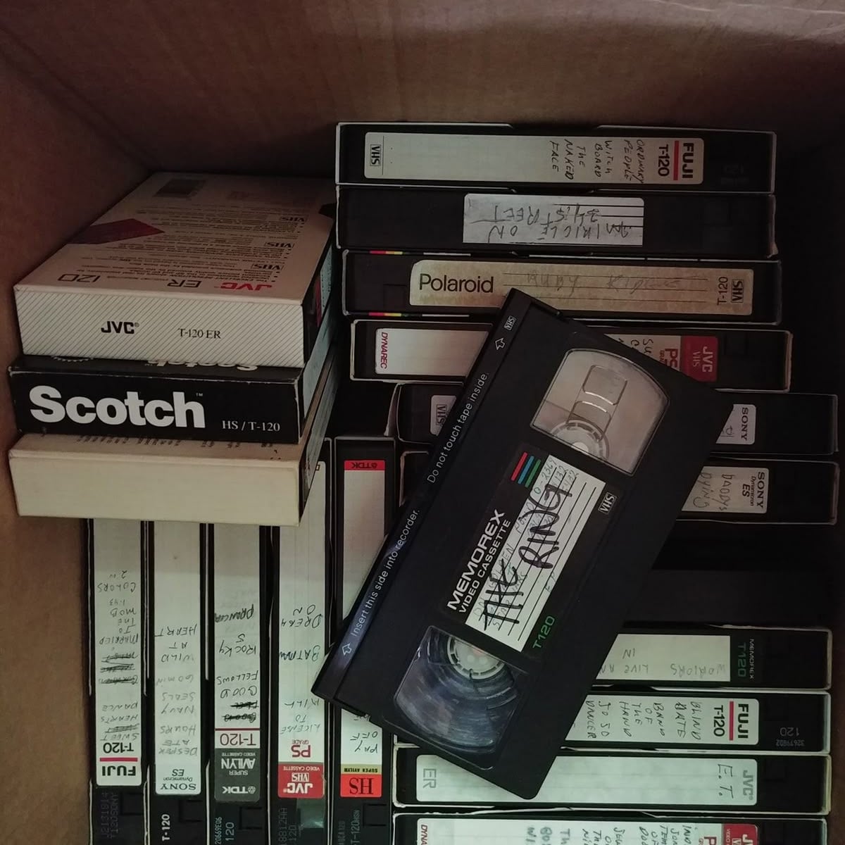 A cardboard box filled with various VHS tapes, some labeled with handwritten notes and different brands like JVC, Scotch, Fuji, Polaroid, and Memorex. One tape lies diagonally on top of the others.