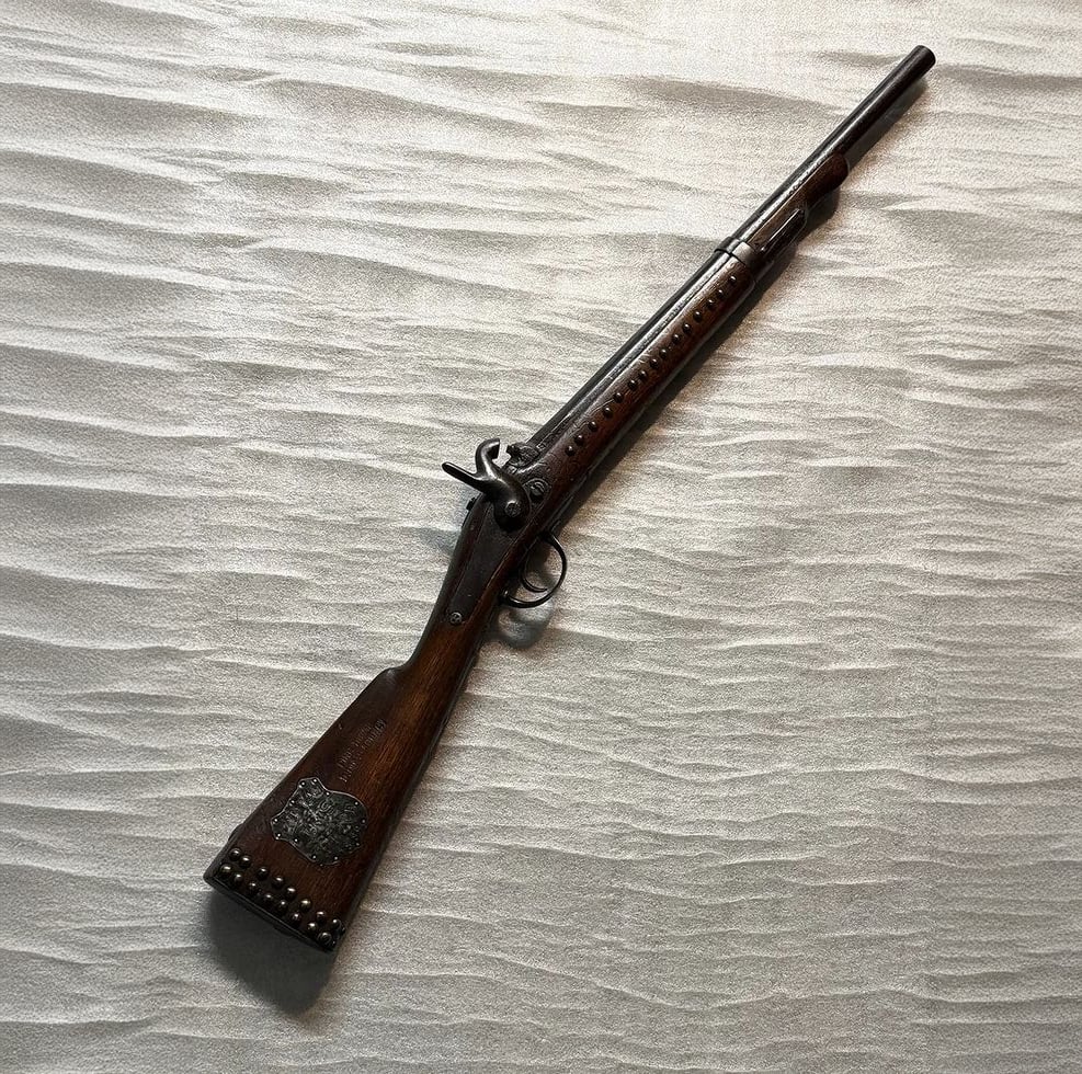 A vintage flintlock musket with a wooden stock and metal accents lies diagonally on a textured, light-colored surface, casting a soft shadow beneath it.