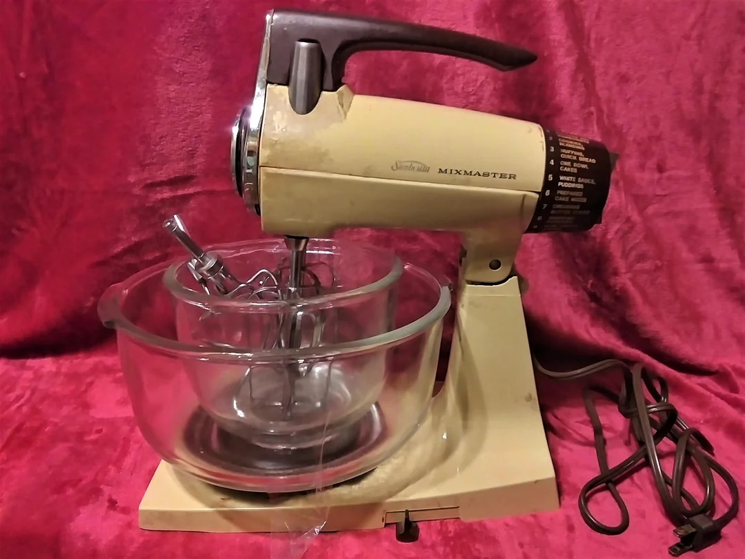 A vintage beige Sunbeam Mixmaster electric stand mixer with a glass mixing bowl and beaters, sitting on a red textured surface with its electrical cord visible.
