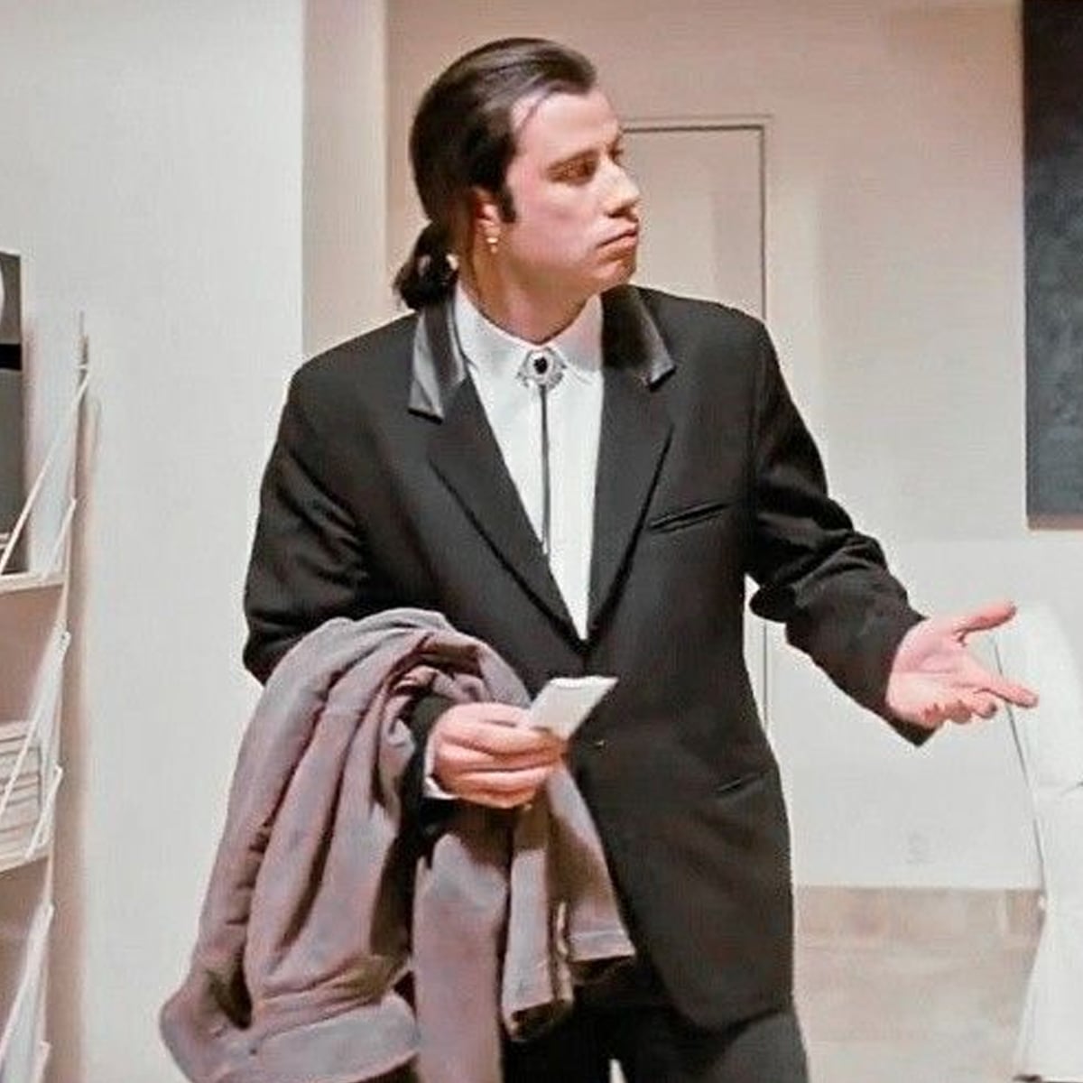 A man with slicked-back hair wearing a black suit and white shirt stands indoors holding a coat over his arm, looking confused with one hand outstretched.