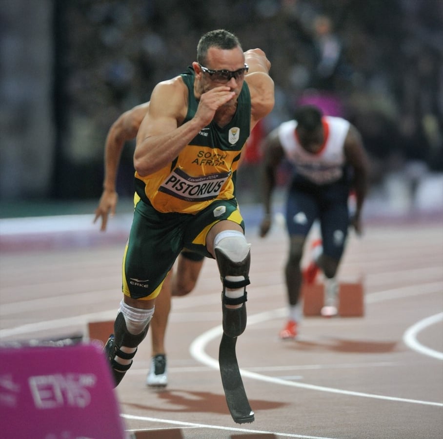 A male Paralympic sprinter wearing prosthetic blades and a green and yellow South African uniform sprints on a track, with competitors visible behind him.