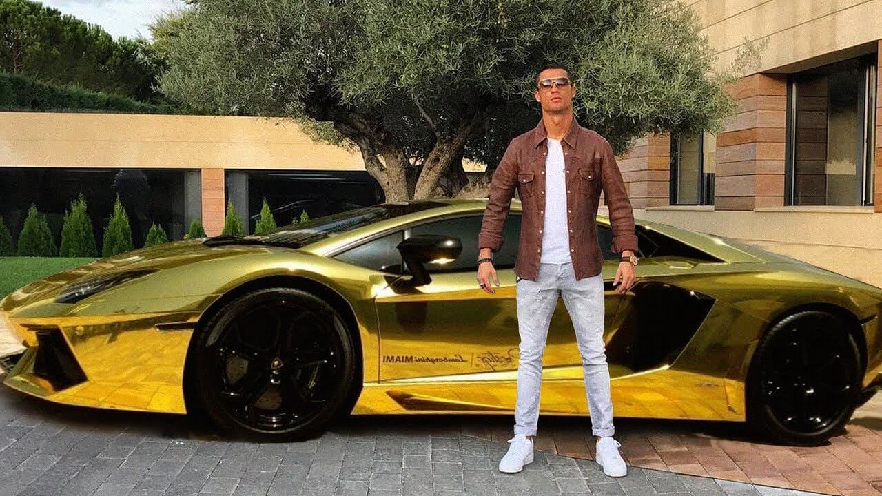 A man in sunglasses, a brown jacket, and white pants stands confidently in front of a shiny gold sports car parked on a paved driveway near a modern building and trees.